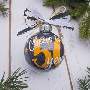 May include: Glittery black Christmas ornament with the text "Cheers to 50 years" in white and orange. The ornament is decorated with a white, black, and gold ribbon bow. Small gold stars are scattered around the number 50.