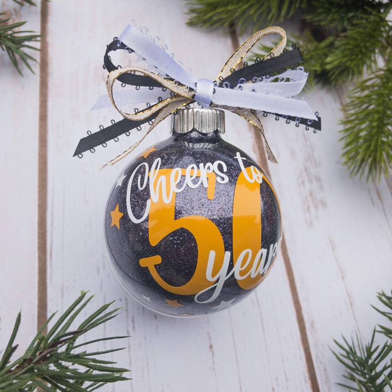 May include: Glittery black Christmas ornament with the text "Cheers to 50 years" in white and orange. The ornament is decorated with a white, black, and gold ribbon bow. Small gold stars are scattered around the number 50.