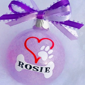 May include: Purple glitter Christmas ornament with a red heart and white paw print, and a white bone with the name "ROSIE" on it. The ornament is topped with a purple and white ribbon.