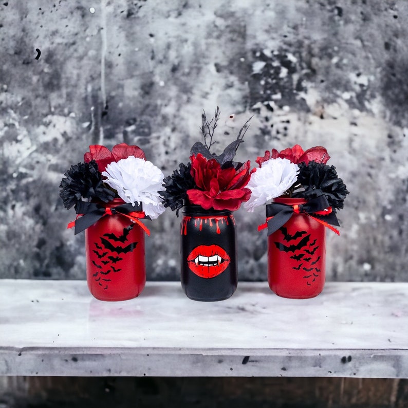 May include: Three Halloween-themed mason jars with floral arrangements. Two red jars feature black bat silhouettes, while the center black jar has a vampire lip design. Each jar holds red, black, and white flowers, tied with black ribbons.