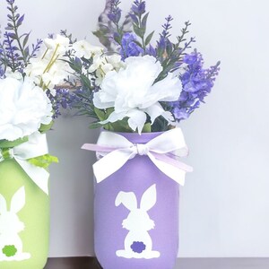 May include: Two mason jars painted in pastel colors, green and purple, with white bunny silhouettes. The jars are filled with white carnations and purple lavender.