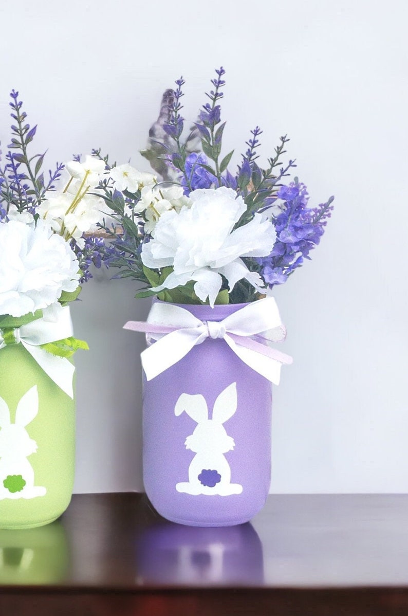 May include: Two mason jars painted in pastel colors, green and purple, with white bunny silhouettes. The jars are filled with white carnations and purple lavender.