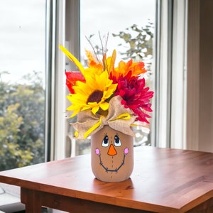 Peut inclure: Un pot Mason peint avec un visage d'&eacute;pouvantail et un arrangement floral d'automne. Les fleurs sont jaunes, rouges et orange.