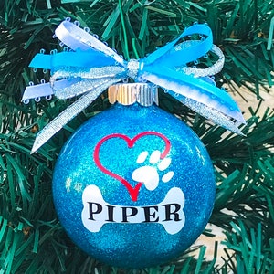 May include: Blue glitter Christmas ornament with a white bone, red heart, and paw print. The bone has the name "PIPER" written on it.