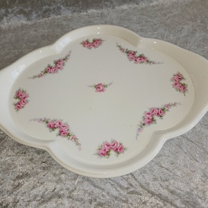 Op de afbeelding: Een wit serveerblad met geschulpte randen en een rozenpatroon. De lade heeft clusters van roze rozen en groene bladeren, met een enkele roos in het midden. De lade is waarschijnlijk gemaakt van keramiek of porselein.