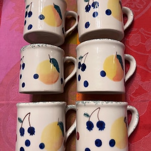 May include: Six white ceramic mugs with a hand-painted design of yellow fruit, blue berries, and green leaves. The mugs have a slightly textured rim and a curved handle. The mugs are arranged on a red and pink patterned tablecloth.