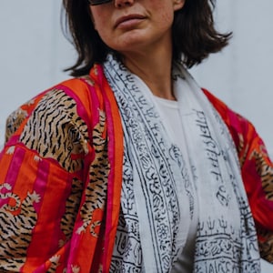 May include: A vibrant kimono-style jacket with a tiger print in shades of orange, pink, and tan. A white scarf with black script is draped over a white top. The jacket has a bold, eye-catching design.