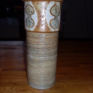 Puede incluir: Un jarrón de cerámica marrón y blanco con una superficie texturizada y un diseño geométrico. El jarrón es alto y cilíndrico.