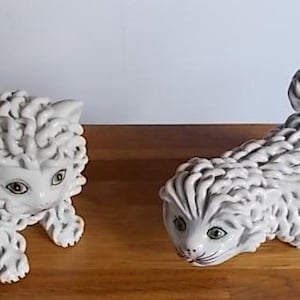 May include: Two white ceramic cat ornaments. One is seated, the other is stretched out with a curled tail. Both have detailed fur, green eyes, and pink noses. The ornaments are displayed on a wooden surface.