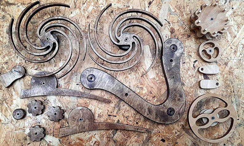May include: A set of wooden gears and intricate wooden pieces, likely for a kinetic sculpture or art project. The pieces are arranged on a wood surface.