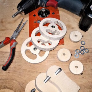 May include: A set of wooden parts for a woodworking project, including four round pieces with a center hole, two curved pieces with a hole, and a set of metal washers. The parts are laid out on a wooden surface with a drill and pliers.