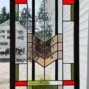 May include: A rectangular stained glass window with a dark brown wooden frame. The design features geometric shapes in clear, red, green, and tan glass. The window is hanging in front of a blurred outdoor scene.