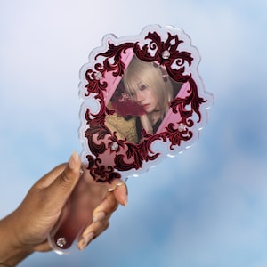 May include: A handheld mirror featuring a clear acrylic frame with a decorative dark red and black filigree border. The mirror surface displays a printed image of a person with blonde hair. Small clear circular accents are embedded in the frame.