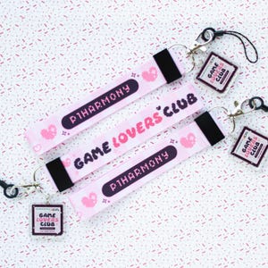 May include: Pink lanyards with the text "P1HARMONY" and "GAME LOVERS CLUB" in a pixelated font. Each lanyard has a black strap and a silver clasp. Small heart and star graphics are also present. Clear charms with the text "GAME LOVERS CLUB" are attached.