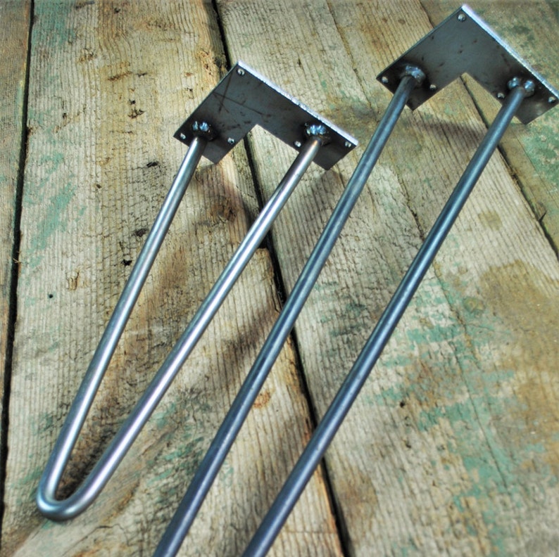 May include: Two metal hairpin legs for furniture, one longer than the other, with a square metal plate at the top. The legs are silver in color and are shown against a wooden background.