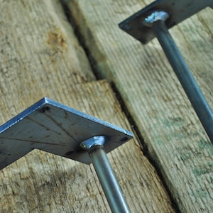 May include: Two metal table legs with square plates. The legs are made of steel and have a black finish. The plates are welded to the legs and have a slightly textured surface.