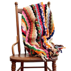 May include: A colorful crocheted blanket with a wavy pattern in shades of pink, orange, green, blue, and brown, draped over a wooden chair.