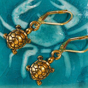 May include: A pair of gold-tone earrings featuring a turtle design. The turtles are detailed with a textured shell and have a hook closure.