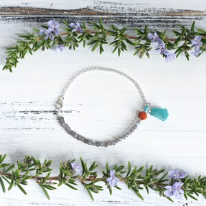Puede incluir: Un brazalete de cadena de plata con una hebra de piedras preciosas grises y un dije de borla turquesa. El brazalete est&aacute; sobre un fondo de madera blanca con ramitas de romero.