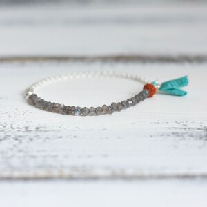 Puede incluir: Un brazalete de cadena de plata con cuentas de labradorita grises y un borla turquesa y naranja.