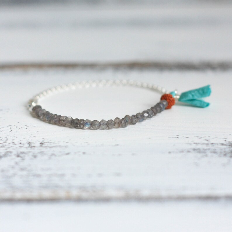Puede incluir: Un brazalete de cadena de plata con cuentas de labradorita grises y un borla turquesa y naranja.