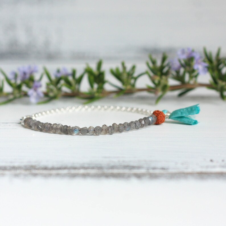 Puede incluir: Un brazalete de cadena de plata con una fila de cuentas de labradorita grises y un borla turquesa con una cuenta naranja.