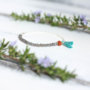 Puede incluir: Un brazalete de cadena de plata con cuentas grises y una peque&ntilde;a cuenta naranja. Un borla turquesa cuelga del brazalete.