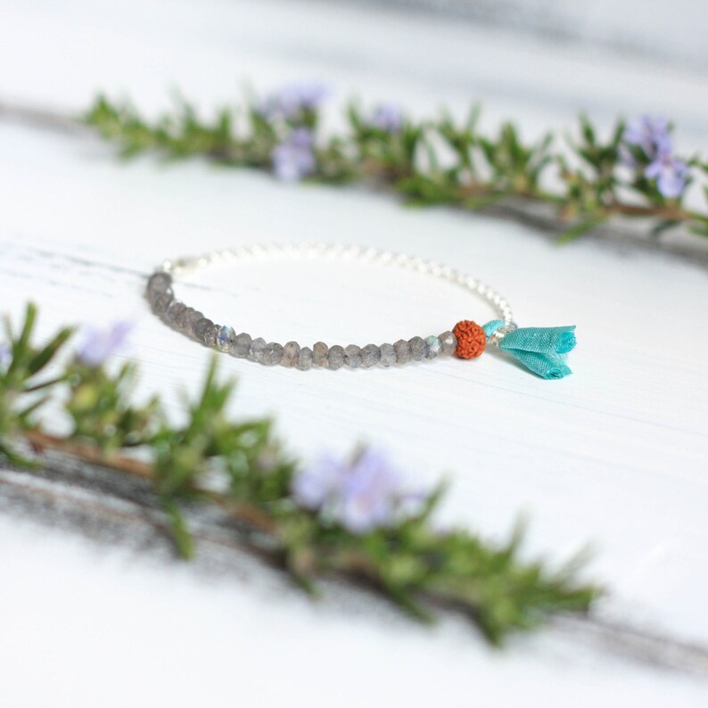 Puede incluir: Un brazalete de cadena de plata con cuentas grises y una peque&ntilde;a cuenta naranja. Un borla turquesa cuelga del brazalete.