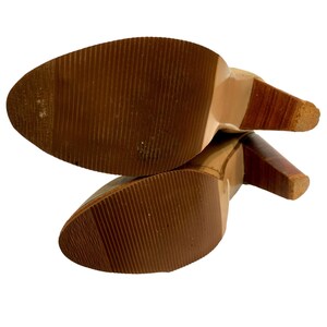 May include: A pair of high-heeled shoes, viewed from the soles. The soles are brown with a ridged texture. The heels are a combination of light and dark brown wood. The shoes are set against a white background.