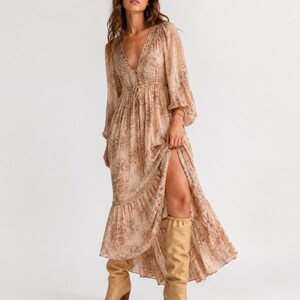 May include: A long, flowing dress with a V-neckline and long sleeves. The dress features a floral pattern in shades of beige and brown. The dress has a high slit and is paired with knee-high tan boots with a heel.