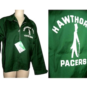 May include: A dark green zip-up jacket with white lettering and a graphic on the back. The jacket features the words "HAWTHORNE AREA YOUTH BAND" and "PACERS". A tag hangs from the zipper. The jacket is displayed on a mannequin.