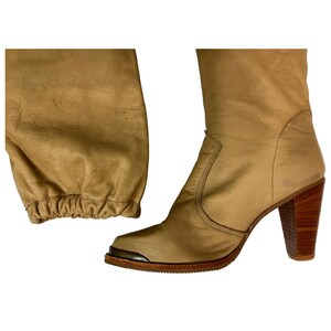 May include: Tan leather knee-high boots with a pointed toe and a wooden block heel. The boots have a metal accent on the toe and a gathered detail at the top of the boot shaft. The heel measures approximately 3 inches.