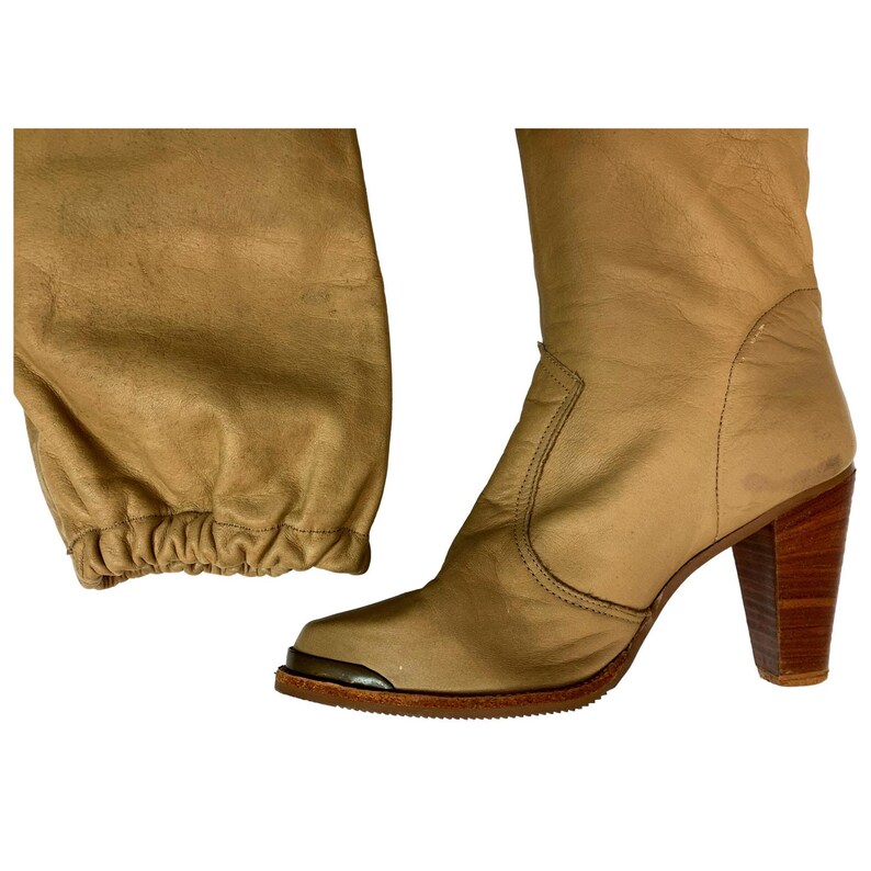 May include: Tan leather knee-high boots with a pointed toe and a wooden block heel. The boots have a metal accent on the toe and a gathered detail at the top of the boot shaft. The heel measures approximately 3 inches.