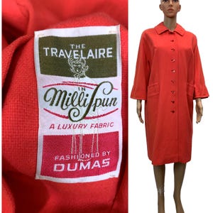 May include: A vintage coral-red coat with a button-down front, three-quarter sleeves, and two front pockets. The coat is displayed on a mannequin. A label reads "The Travelaire Milli spun A Luxury Fabric Fashioned by Dumas."
