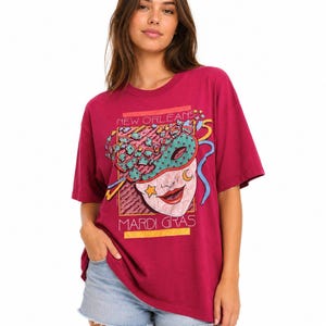 May include: A magenta short-sleeve t-shirt featuring a graphic print of a teal masquerade mask with a yellow star and crescent moon. The text "NEW ORLEANS" appears at the top and "MARDI GRAS" at the bottom of the rectangular design.