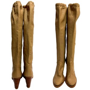 May include: A pair of knee-high tan leather boots. The boots have a rounded toe, a wooden heel, and a gathered detail at the top. The boots are displayed on a white background.