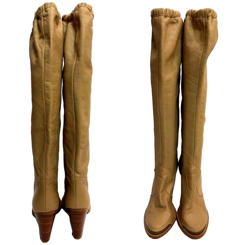 May include: A pair of knee-high tan leather boots. The boots have a rounded toe, a wooden heel, and a gathered detail at the top. The boots are displayed on a white background.
