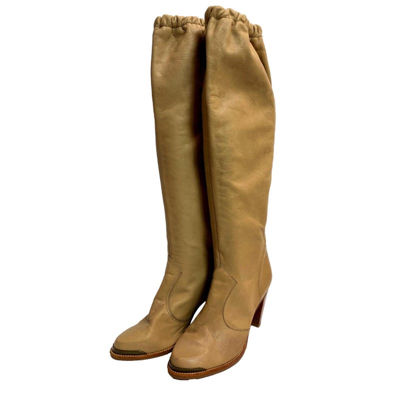 May include: A pair of knee-high tan leather boots with a rounded toe and a stacked heel. The boots have a gathered top and a subtle sheen, suggesting a vintage style. The heels are approximately 3 inches high.