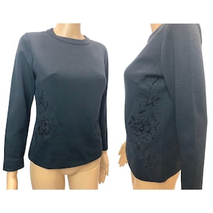 May include: A dark teal long-sleeve top with a crew neck. The top features a floral design on the side, embellished with beads. The design is on a panel that extends from the waist to the shoulder. The top is displayed on a mannequin.