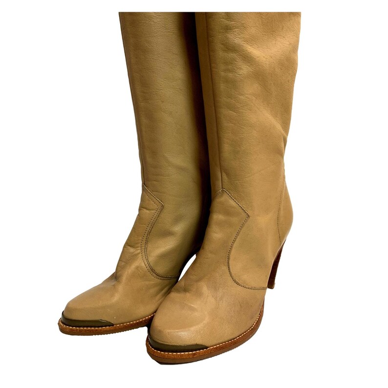 May include: A pair of tan leather knee-high boots. The boots have a pointed toe, a high heel, and a decorative stitch detail along the side. The soles are a darker brown with a gold-tone metal accent.