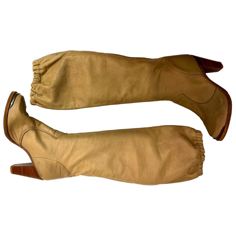 May include: A pair of knee-high tan leather boots. The boots have a rounded toe, wooden heels, and a gathered detail at the top of the boot shaft. The heels are approximately 3 inches (7.6 cm) high.
