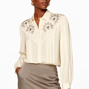 May include: A cream-colored long-sleeve button-down shirt featuring a pointed collar and vertical pleats on the front. The shoulder panels are decorated with intricate embroidery and circular metallic accents. The shirt is paired with a taupe skirt.
