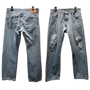 Könnte beinhalten: Hellblaue Jeans aus Denim mit Used-Details, vorne und hinten gezeigt. Merkmale sind Knopfleiste, klassisches Fünf-Taschen-Design und ein Lederpatch. Die Jeans hängen an einem schwarzen Kleiderbügel.