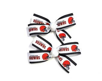 May include: Two white hair bows with black trim and the words "Cleveland Browns" printed on them. Each bow features a red football helmet graphic. The bows are tied with white ribbon.