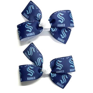 May include: Two navy blue hair bows with a white center. The bows are decorated with the Seattle Kraken logo and the word "KRAKEN" in white. The bows are on a white background.