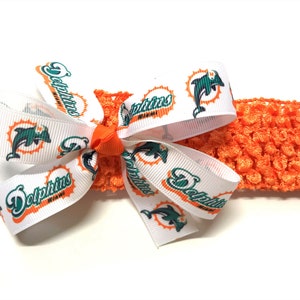 May include: An orange crocheted headband with a white ribbon bow featuring the Miami Dolphins logo. The bow is accented with an orange center. The headband is a sports-themed accessory.