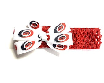 May include: A red crocheted headband with a white bow featuring a repeating pattern of black, red, and white oval designs. The bow is centered on the headband, creating a decorative accessory.