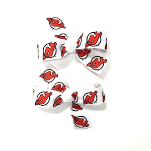 May include: Two white hair bows with a repeating pattern of the New Jersey Devils logo in red and black. The bows are tied and have a classic bow shape, perfect for a fan.