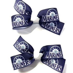 May include: Two navy blue hair bows with the word "MARINERS" printed in white. Each bow features the Mariners logo, a compass rose, and is tied with a white ribbon. The bows are designed for hair accessories.