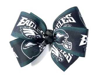 May include: A hair bow in dark green and black, featuring the Philadelphia Eagles logo. The ribbon displays the team's name and helmet in white. The bow is knotted in the centre with a black tie, suitable for a fan.